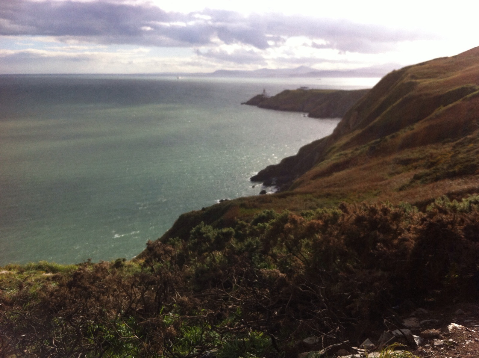 View south along the Howth Head cliff path over Dublin Bay, with the Baily Lighthouse on the headland and the Dublin mountains faint on the horizon